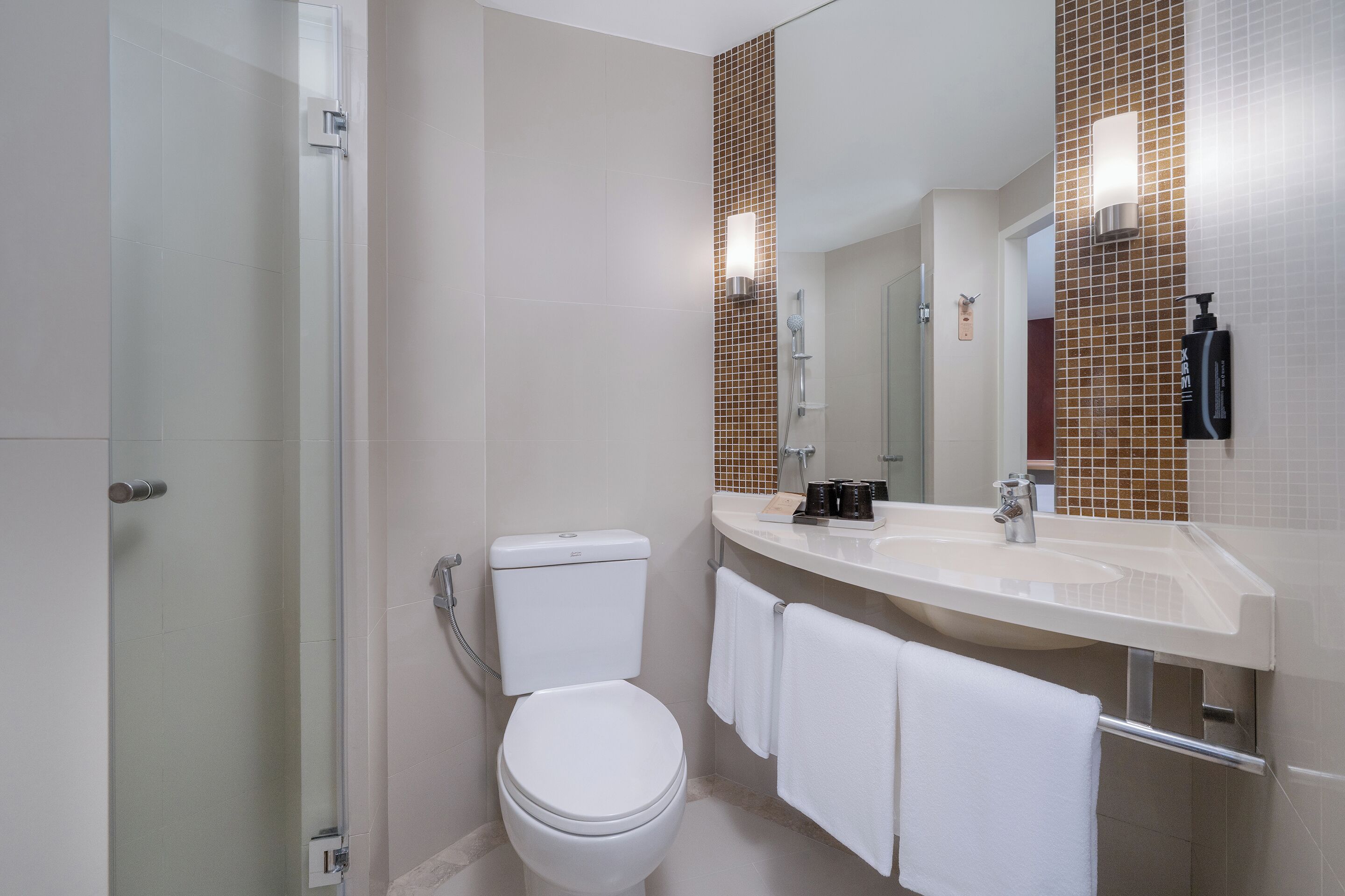 Een moderne badkamer met een douchecabine van glas, een wit toilet, een grote spiegel en een beige aanrecht met een wastafel en toiletartikelen.
