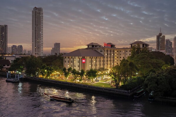 A high-rise hotel with a rooftop terrace, a river in the foreground, and surrounding modern buildings. - ibis Bangkok Riverside (Bangkok)