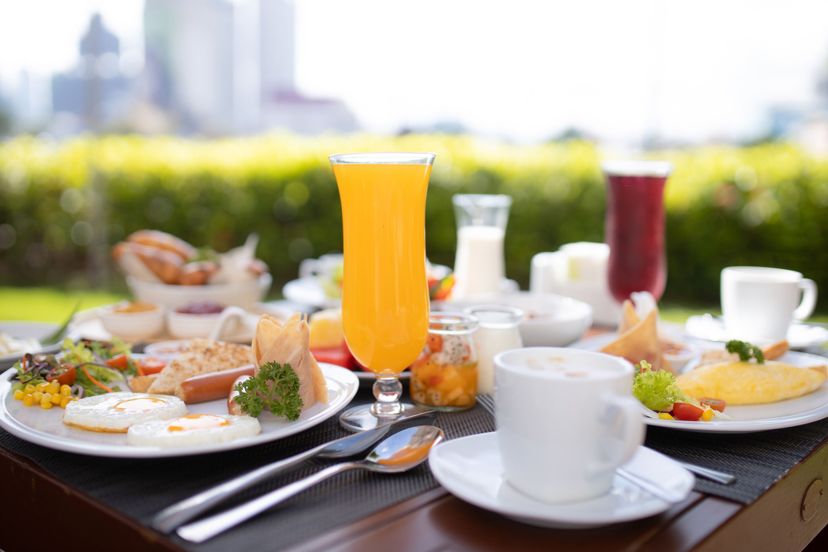 Un bufé de desayuno con diversos platos, un vaso de zumo de naranja y un paisaje urbano al fondo.