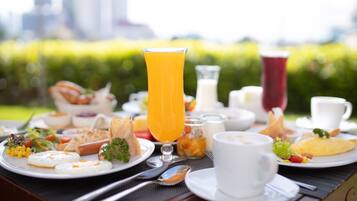 Un bufé de desayuno con diversos platos, un vaso de zumo de naranja y un paisaje urbano al fondo.
