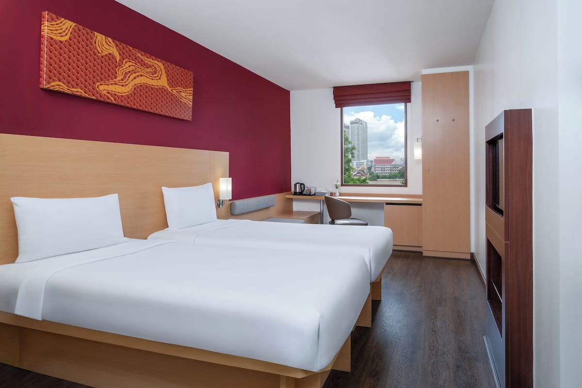 A hotel room with a large bed, a desk, a chair, a TV, and a window with a city view.
