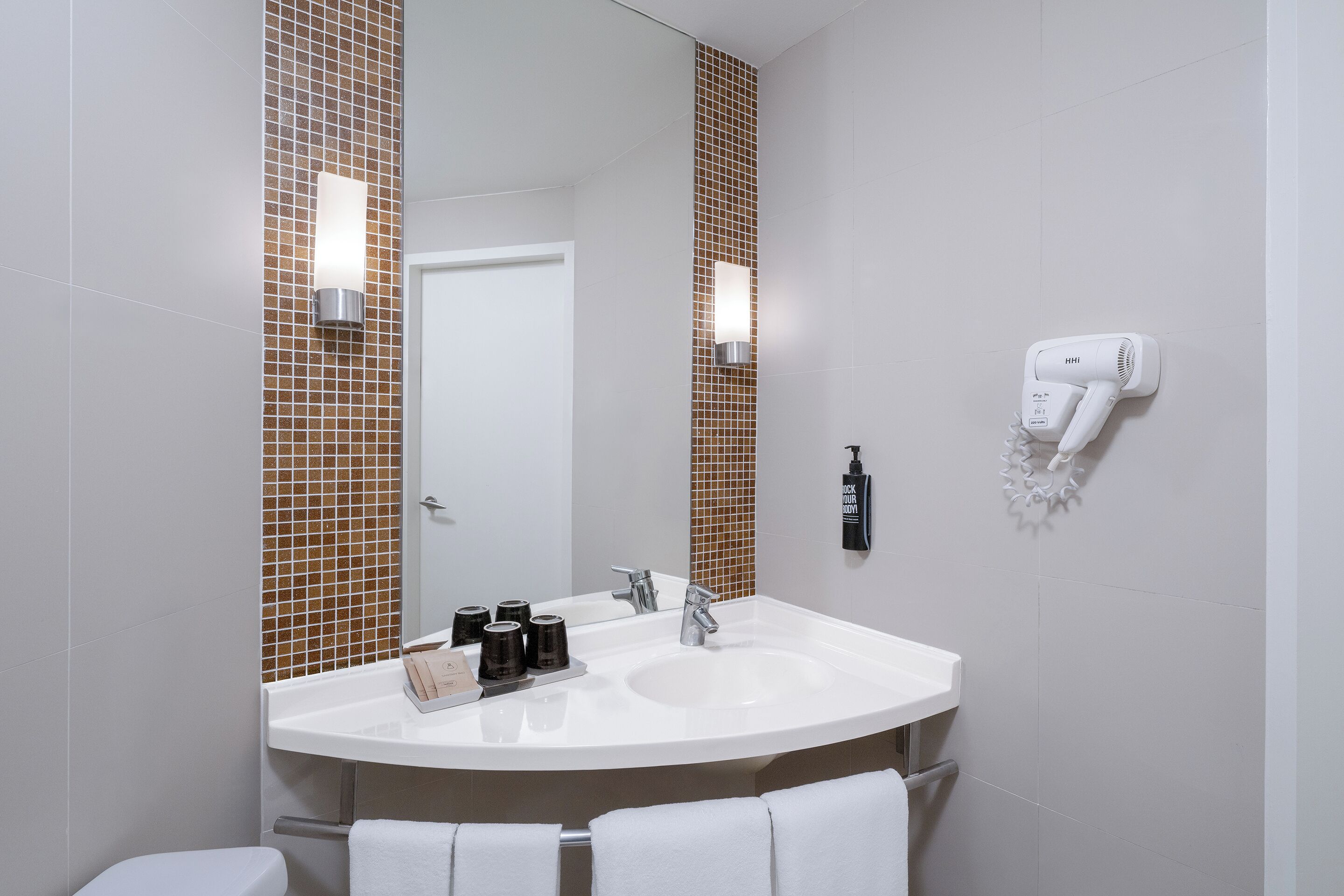 Un baño con un lavamanos blanco, un espejo, un perchero para toallas y un secador de pelo montado en la pared.