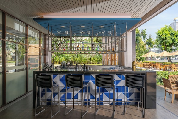 Uma área de bar com parede revestida de azulejos azuis e brancos, uma estrutura de vidro e mesas ao ar livre.