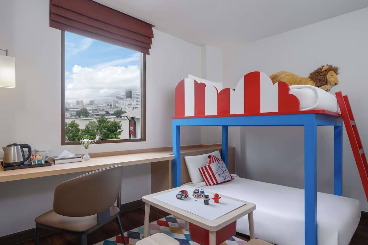 A child's bunk bed with a lion toy, a desk with a laptop, a chair, and a window with a city view.