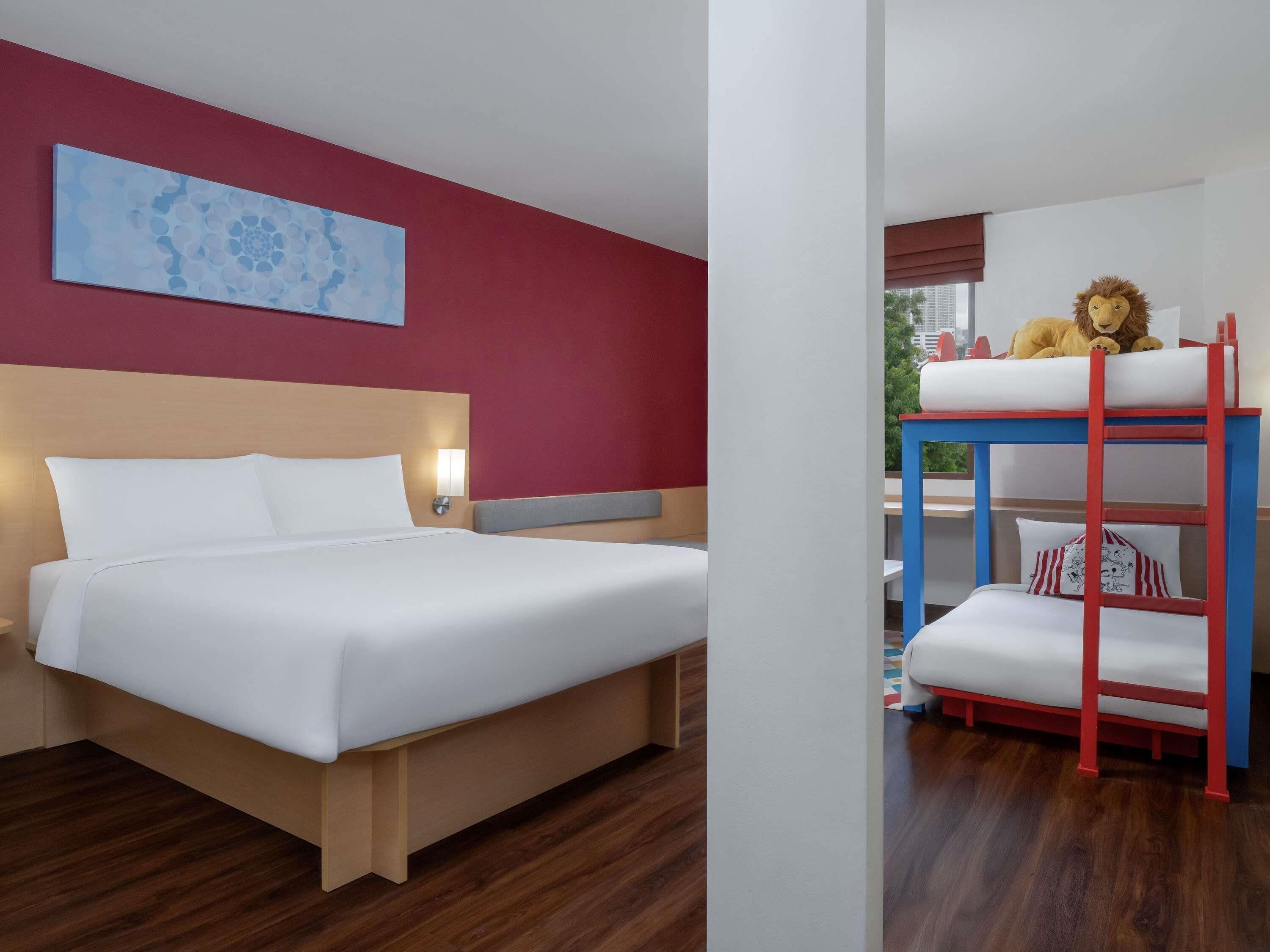 A hotel room with a bed, a bunk bed, a wall art piece, and a window with a view.