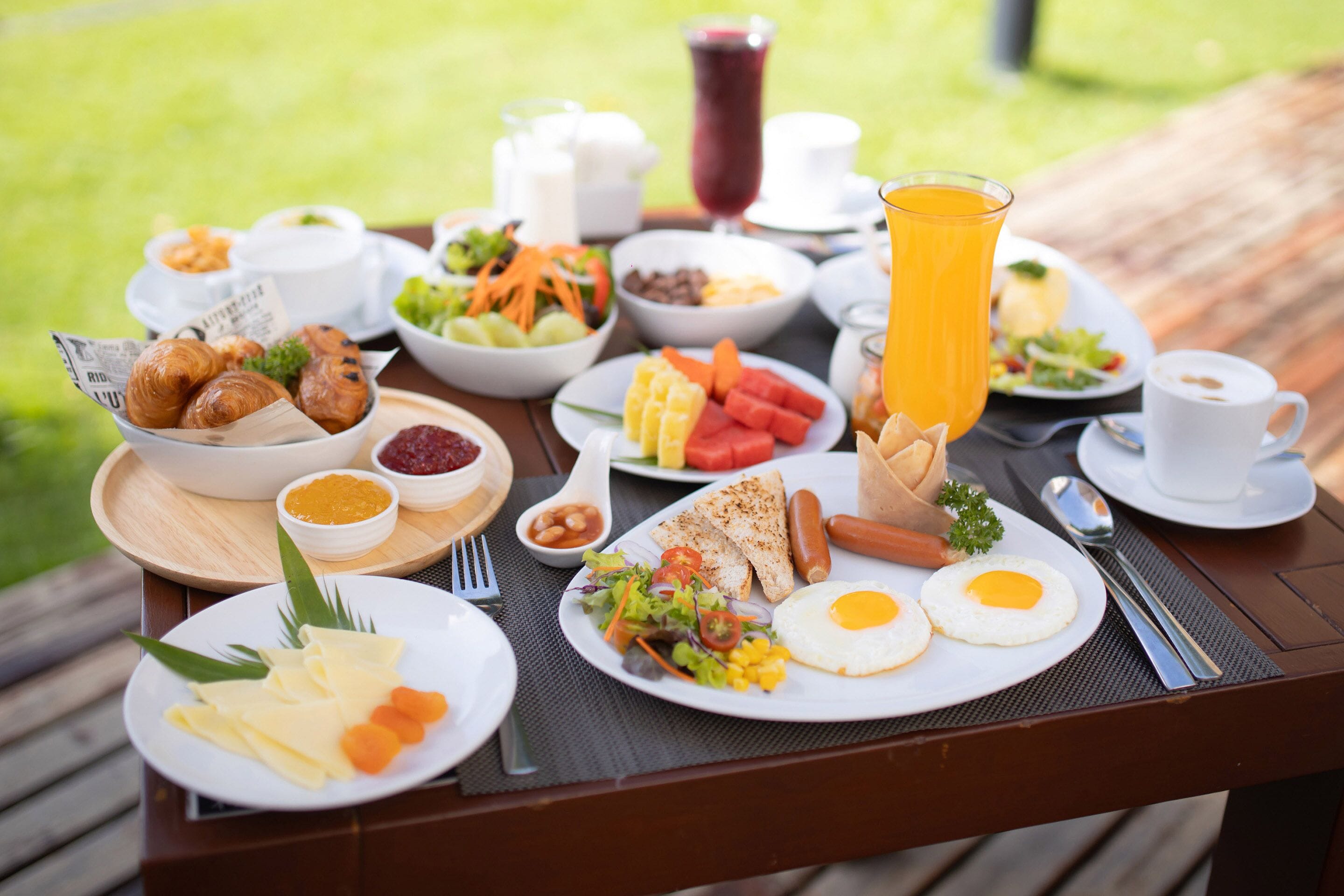 Un bufé de desayuno con diversos platos, entre ellos huevos, salchichas y frutas frescas, sobre una mesa de madera al aire libre.