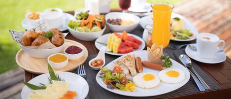 Un bufé de desayuno con diversos platos, entre ellos huevos, salchichas y frutas frescas, sobre una mesa de madera al aire libre.