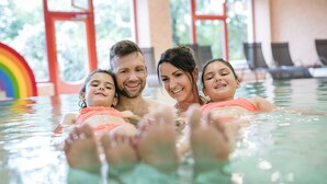 Une famille se trouve dans une piscine intérieure à l’eau claire, décorée d’un arc-en-ciel, et offre une vue sur l’extérieur grâce à de grandes fenêtres.