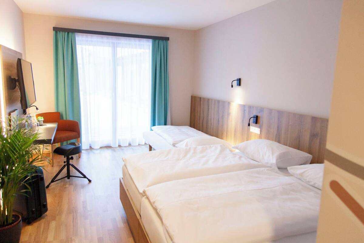 Une chambre d’hôtel avec deux lits, un bureau avec une chaise, une plante en pot et une grande fenêtre avec des rideaux.