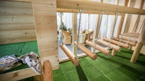Une structure de jeu en bois, dotée de balançoires et d’un petit espace clos recouvert d’une surface verte.