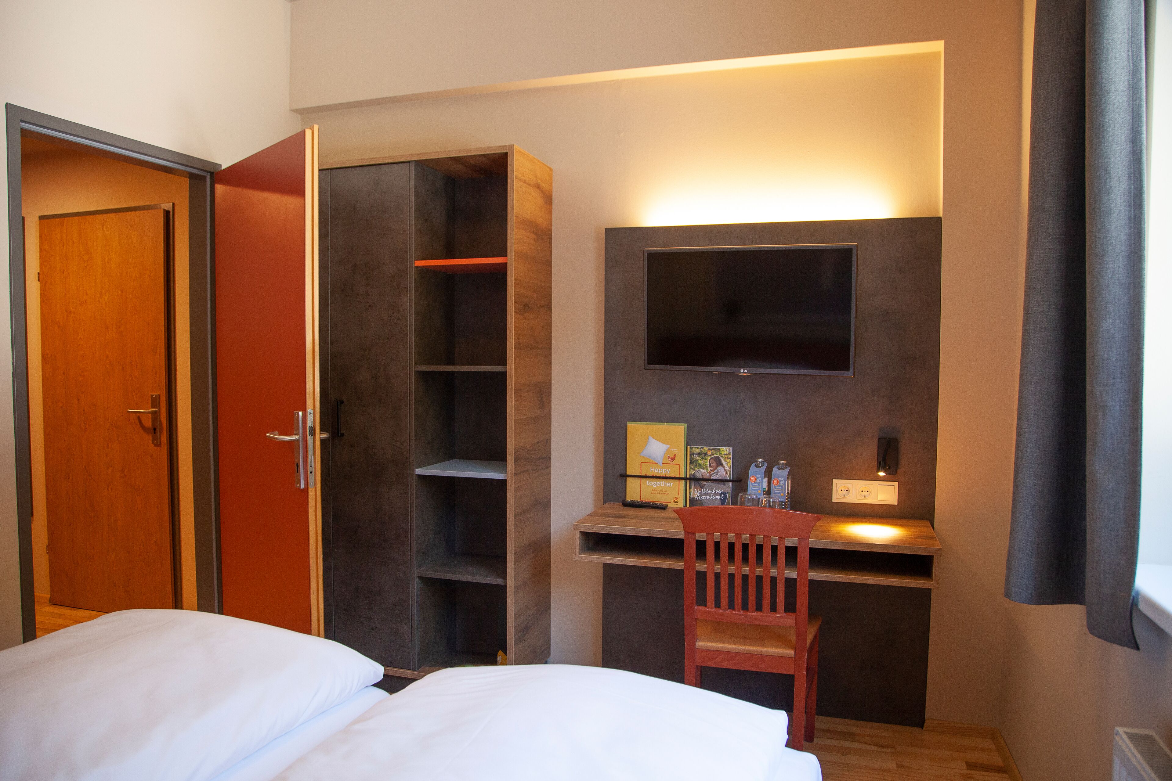 Quarto de hotel com cama, escrivaninha com cadeira, televisão na parede e guarda-roupa.