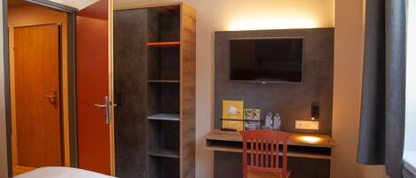A hotel room with a bed, a desk with a chair, a TV mounted on the wall, and a wardrobe.