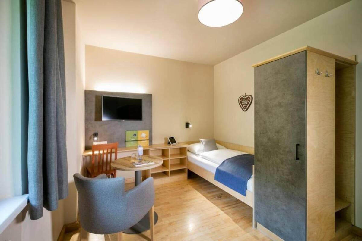 Une chambre d’hôtel de taille réduite, comprenant un lit, un bureau, une chaise, un téléviseur fixé au mur et une armoire.