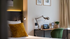 Een hotelkamer met een bed, een bureau met lamp, een groene stoel, een kleine tafel met een plant en ingekaderde foto's aan de muur.