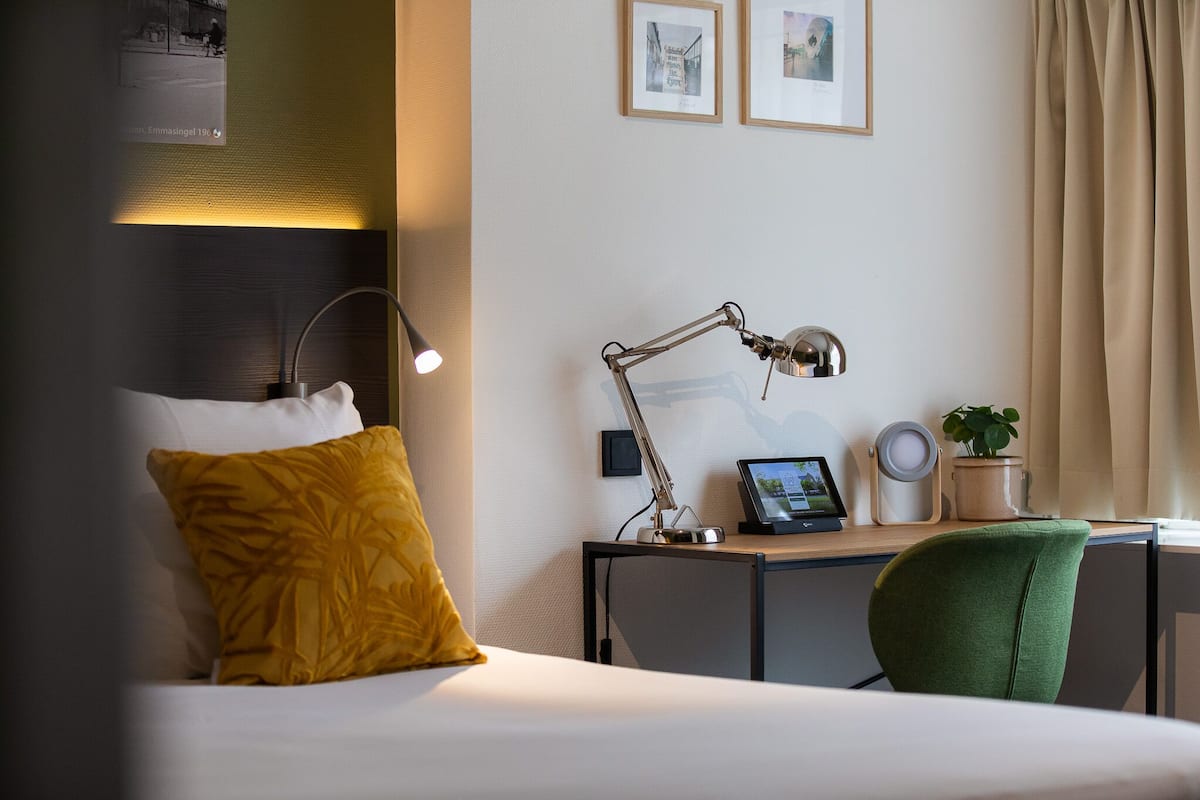 Un cuarto de hotel con una cama, un escritorio con una lámpara, una silla verde, una mesita con una planta y cuadros en la pared.