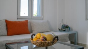 Una cesta de mimbre sobre una mesa de vidrio con frutas, un sofá con un cojín naranja y una ventana al fondo.