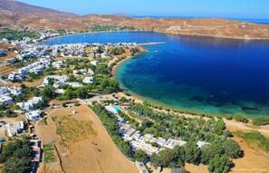 An aerial view of a coastal area with numerous white buildings, a clear blue sea, and a sandy beach. - Indigo Studios (Serifos)