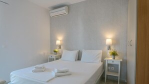 Una cama bien hecha con sábanas y toallas blancas, dos mesitas de noche con lámparas y pequeñas macetas, y un equipo de aire acondicionado en la pared.
