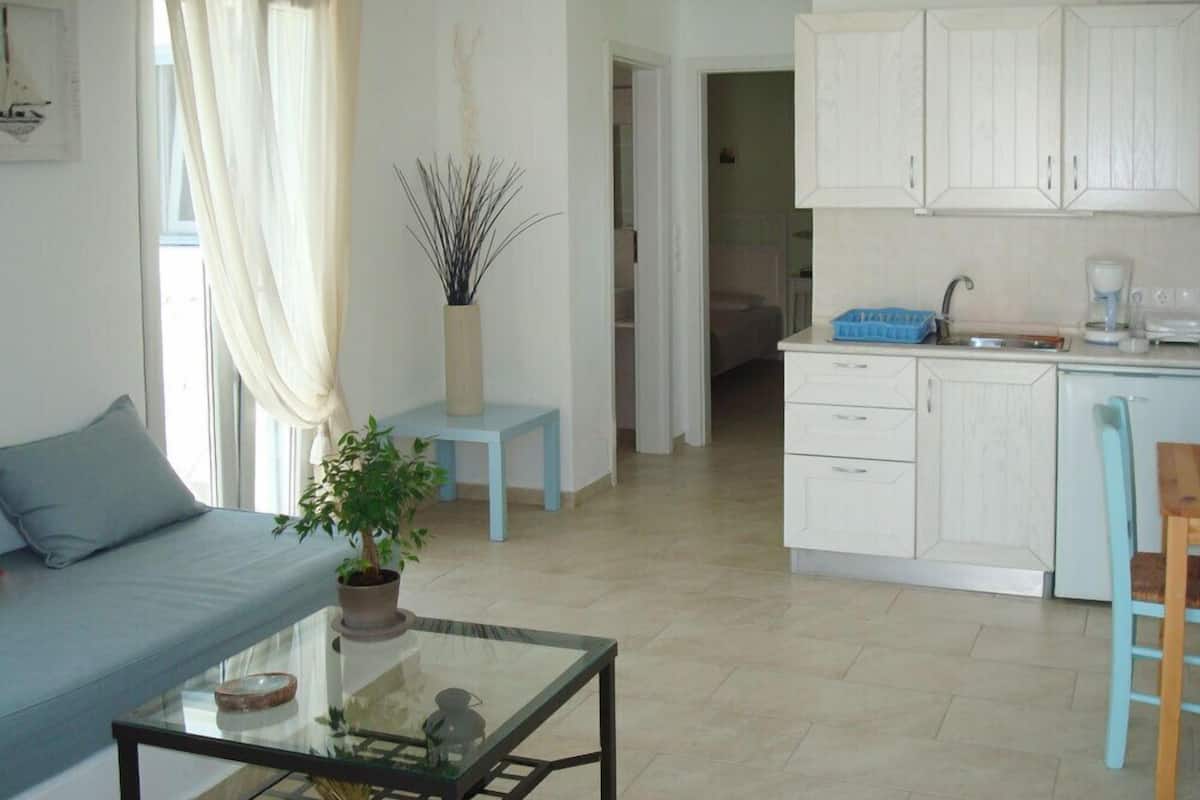 A modern living room with a glass coffee table, a blue sofa, and a potted plant. A kitchen with white cabinets and a blue chair is visible in the background.