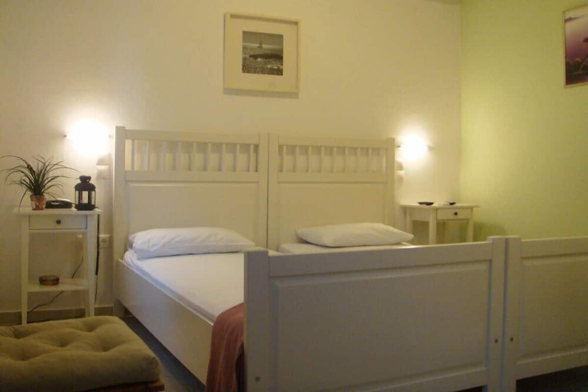 A bedroom with a double bed, bedside tables, a nightstand, a lamp, a potted plant, and an air conditioning unit.
