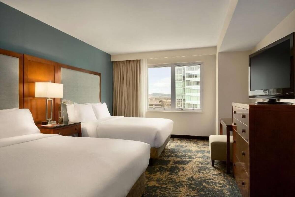 A hotel room with two beds, a television, a dresser, and a window with a city view.