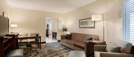 A hotel room with a sofa, armchairs, a desk, and a TV. There is a carpeted floor and a window with blinds.