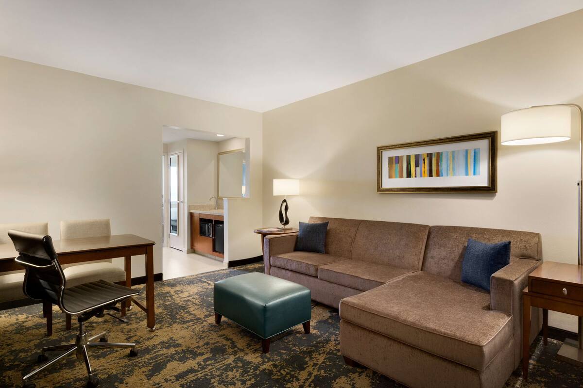 A hotel room with a sofa, ottoman, chair, and a dining table.