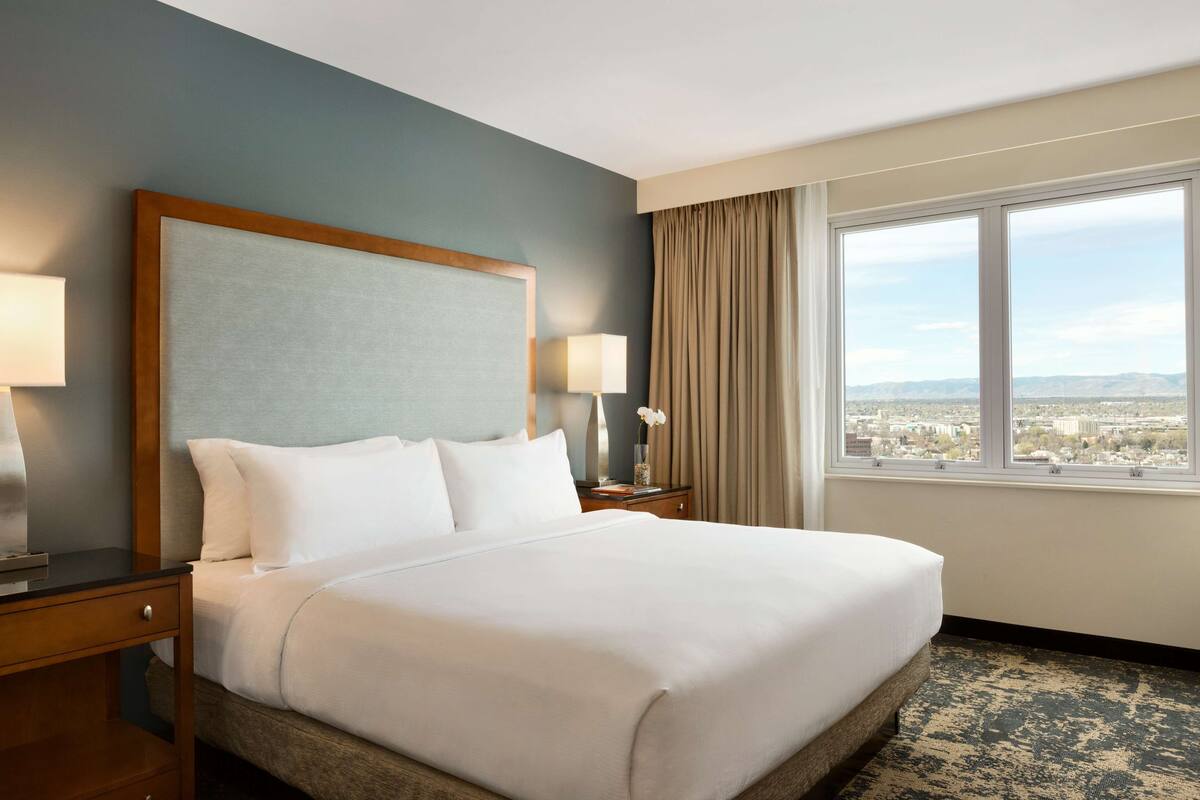 A hotel room with a large bed, two bedside tables with lamps, a view of the city through a large window, and a cityscape visible outside.