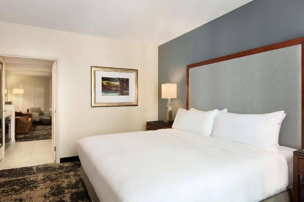 A hotel room with a large bed, a framed picture on the wall, a nightstand with a lamp, and a door leading to a sitting area.