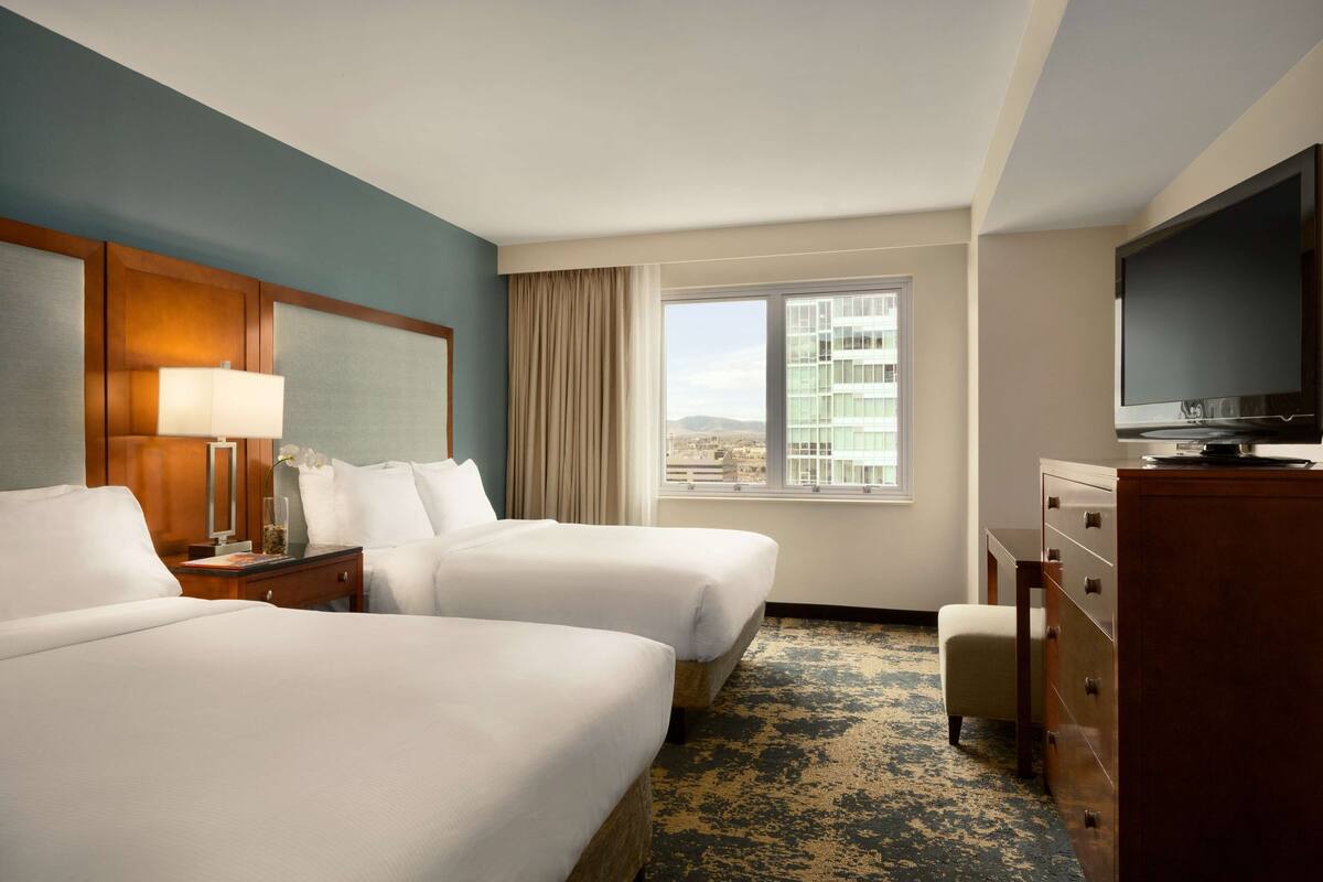 A hotel room with two beds, a television, a dresser, and a window with a city view.