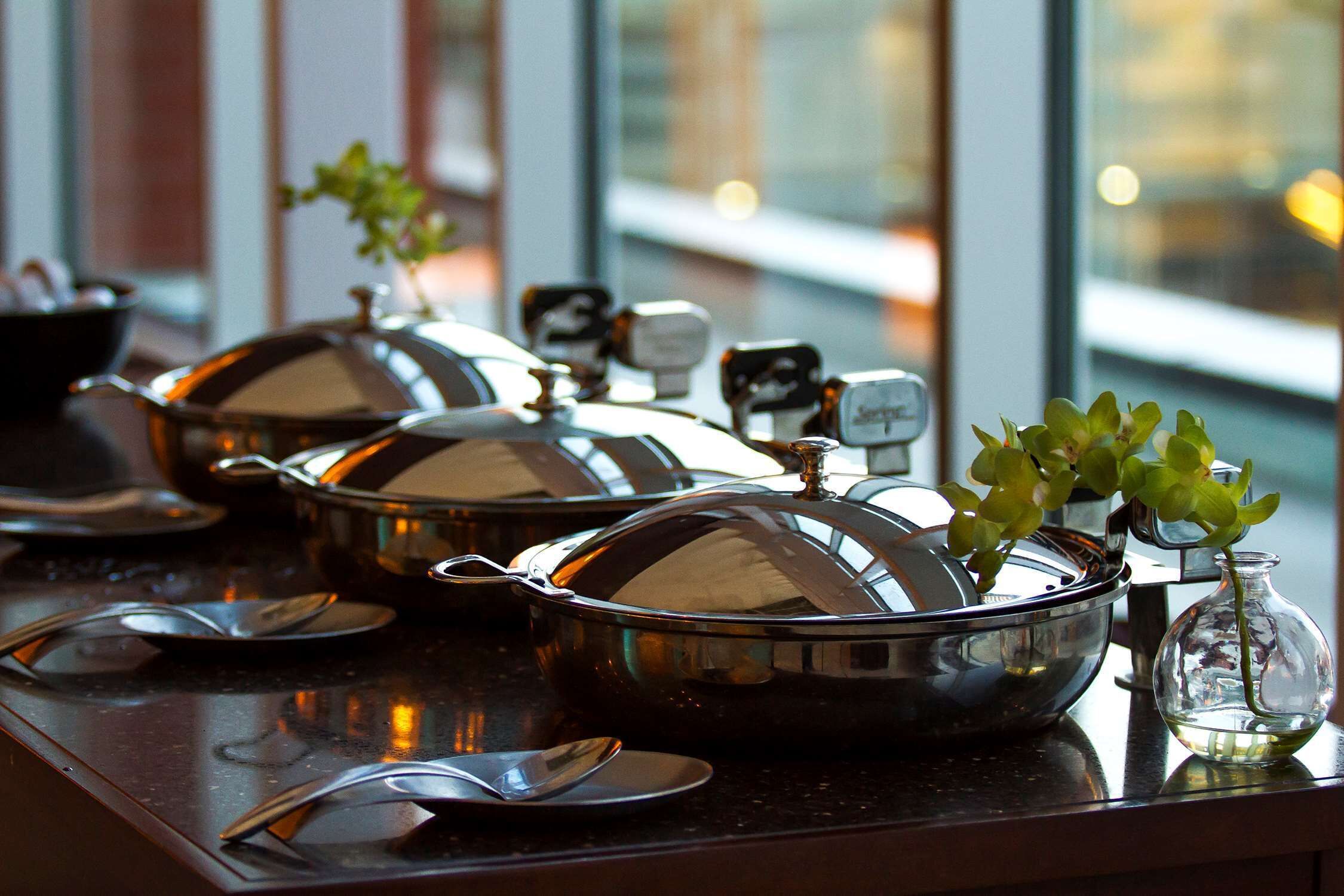 Un espace repas agrémenté de plateaux de buffet en acier inoxydable, d’un vase contenant des plantes vertes et d’une carafe en verre posée sur un comptoir en marbre.