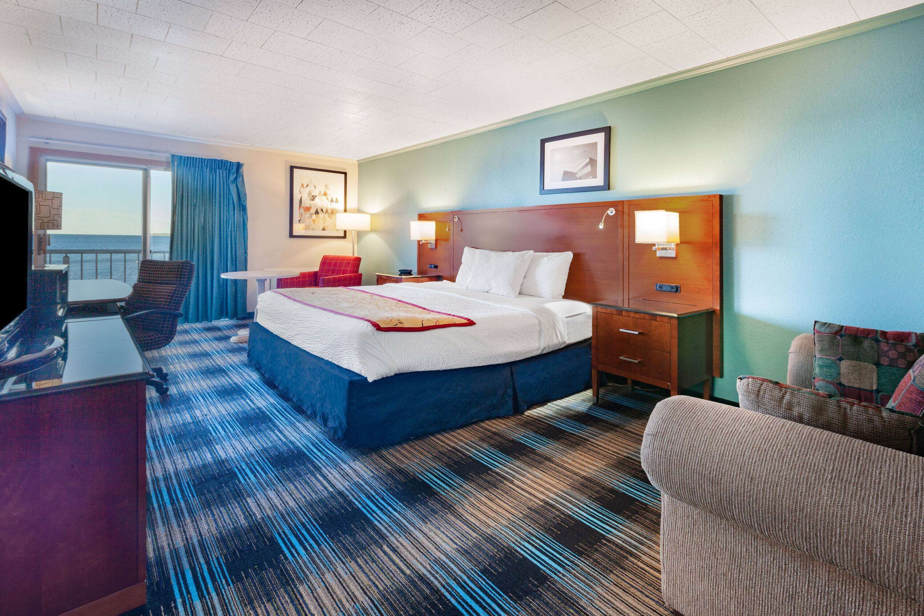 A hotel room with a bed, a desk, a chair, a small table, and a view of the sea through a window.