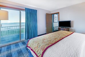 A hotel room with a large bed, a television, a sliding glass door to a balcony, and a view of the sea.