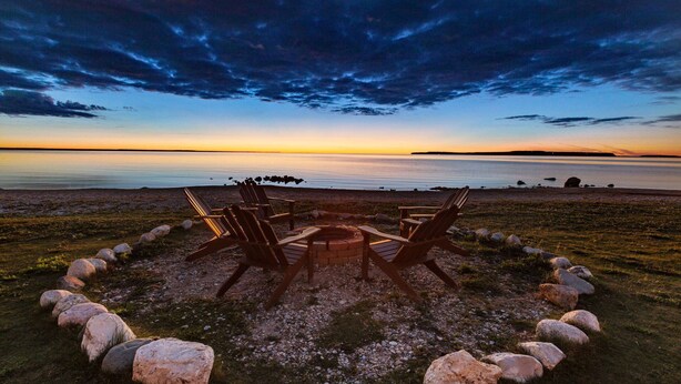 Un espace de détente circulaire, composé de chaises disposées en cercle autour d’un foyer central, le tout entouré de rochers et offrant une vue sur une étendue d’eau calme au coucher du soleil.