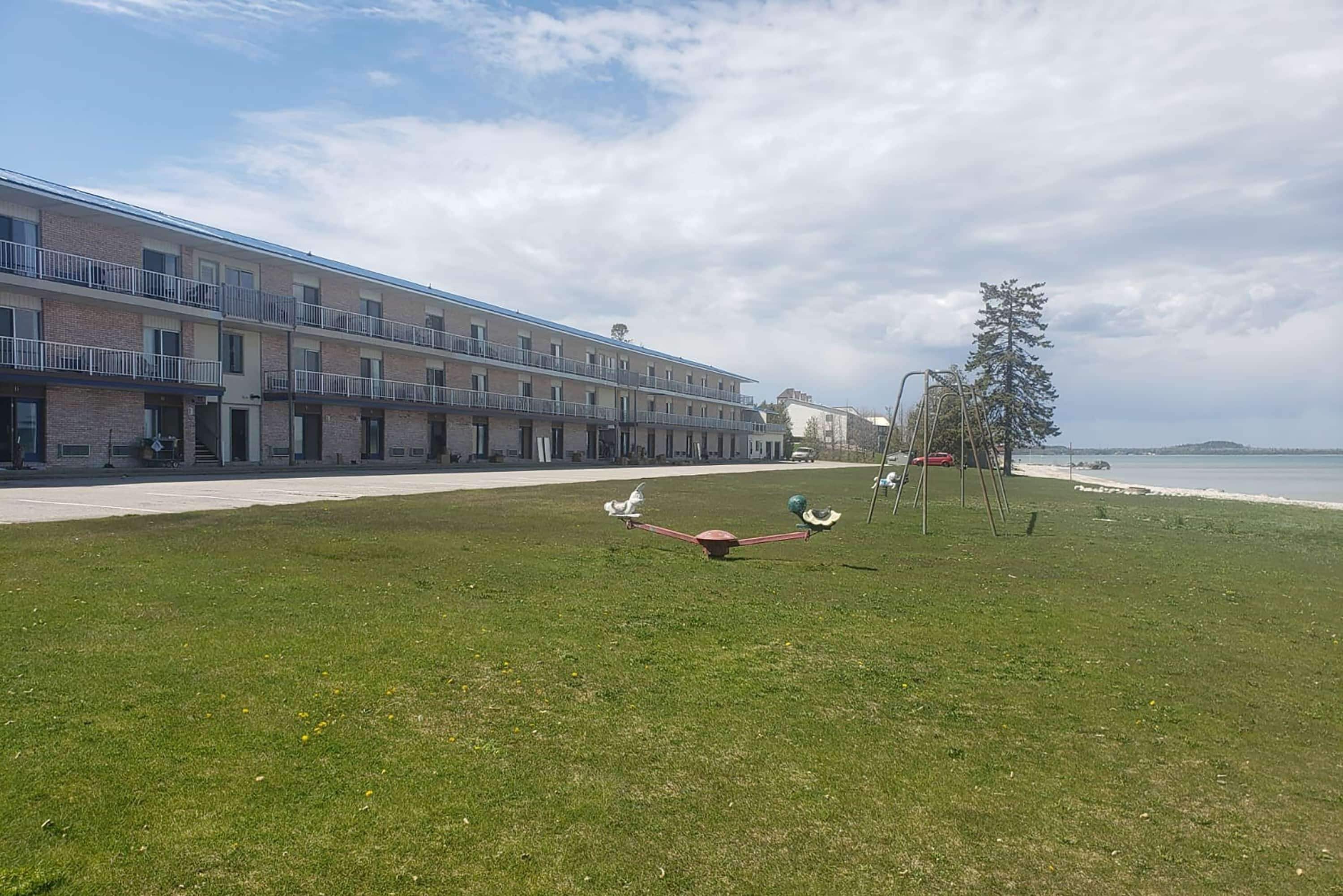A grassy area with playground equipment, a building with balconies, and a beach in the background.