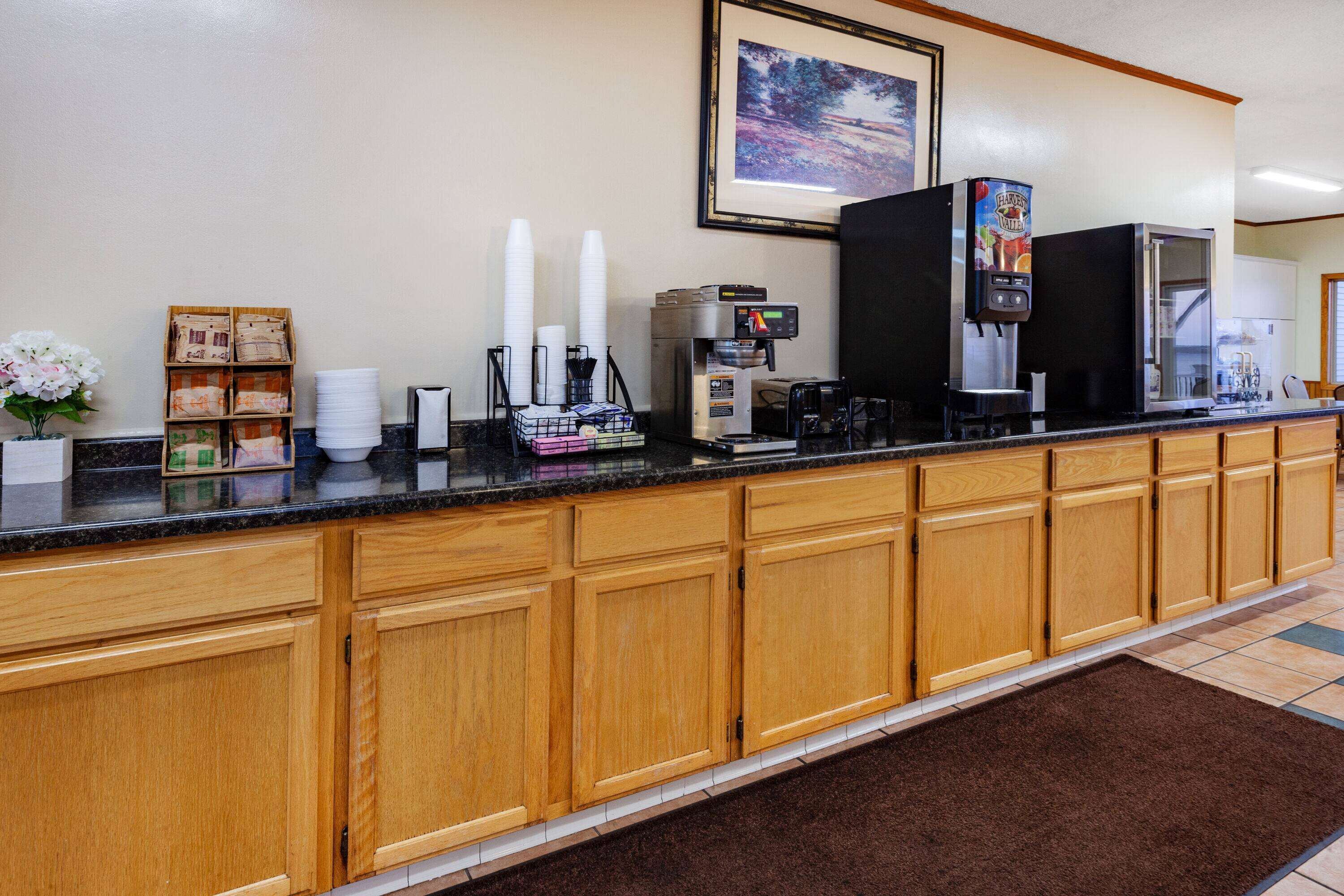 A hotel reception area with a long counter, wooden cabinets, a coffee machine, a beverage dispenser, and a framed picture on the wall.