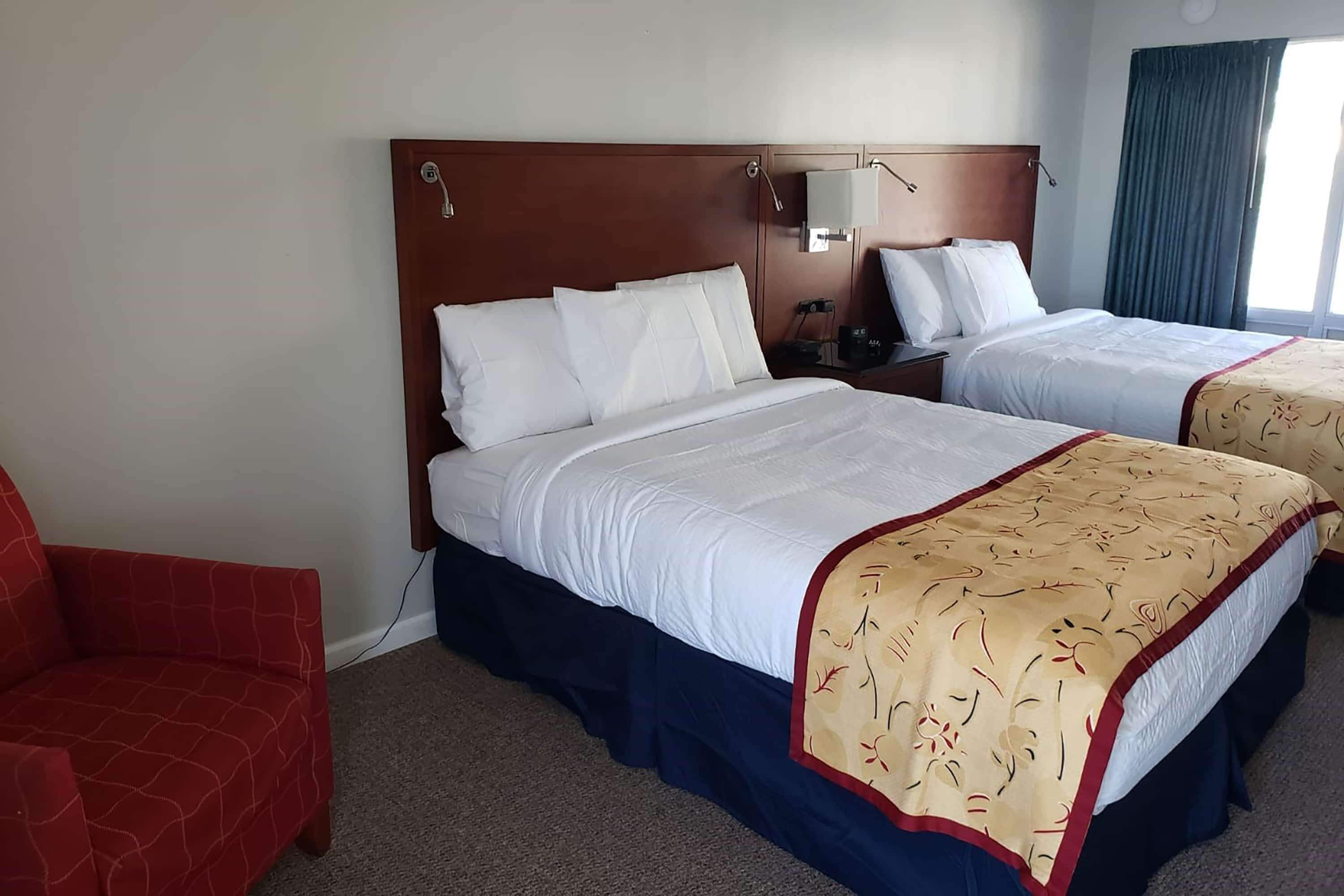 Una habitación de hotel con dos camas, un sillón rojo, un cabecero de madera y una mesita de noche con una lámpara.