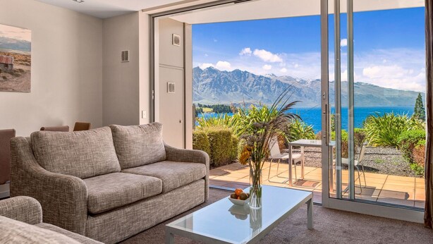 Ein modernes Wohnzimmer mit einer Couch und einem Glastisch, eine Glasschiebetür führt zu einem Balkon mit Blick auf Berge und Meer.
