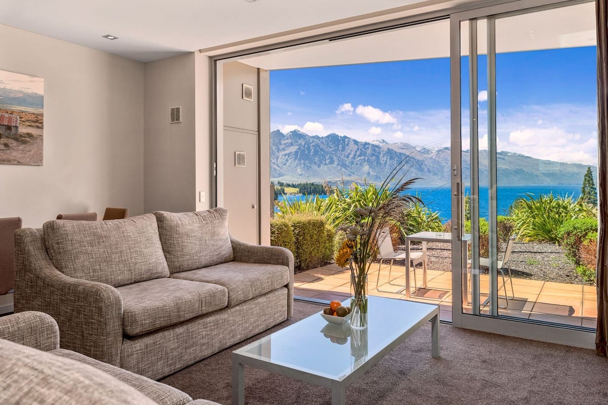 A modern living room with a sofa and a glass coffee table, a sliding glass door leading to a balcony with a mountain and sea view.