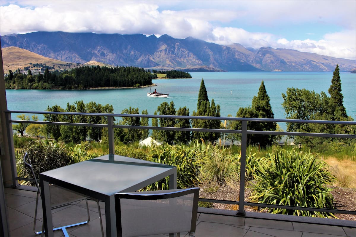 A balcony with a table and chairs overlooking a lake with a boat, surrounded by mountains and greenery.