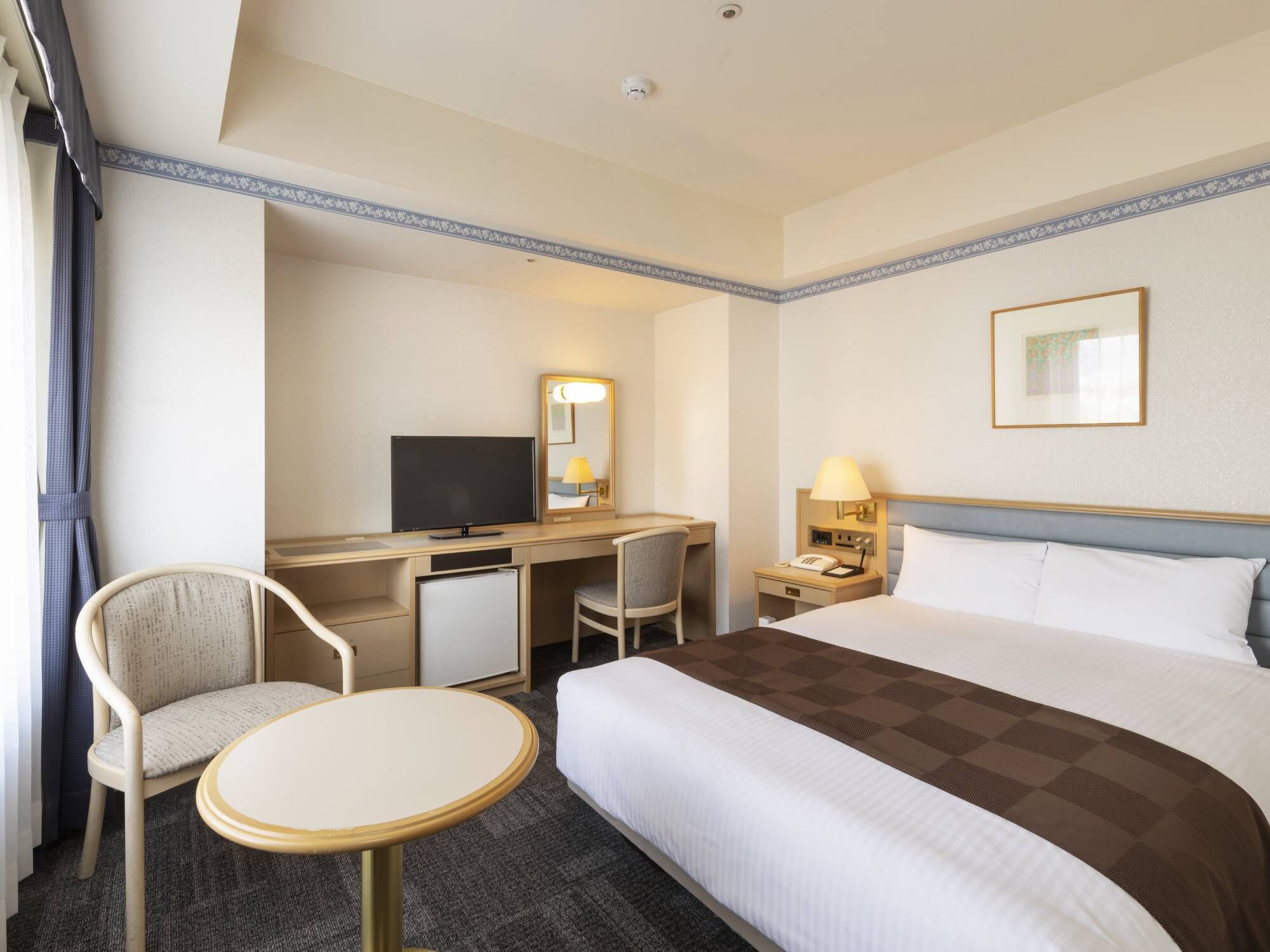 Habitación de hotel con cama, escritorio, silla, televisor, mesita de noche, lámpara y un cuadro en la pared.