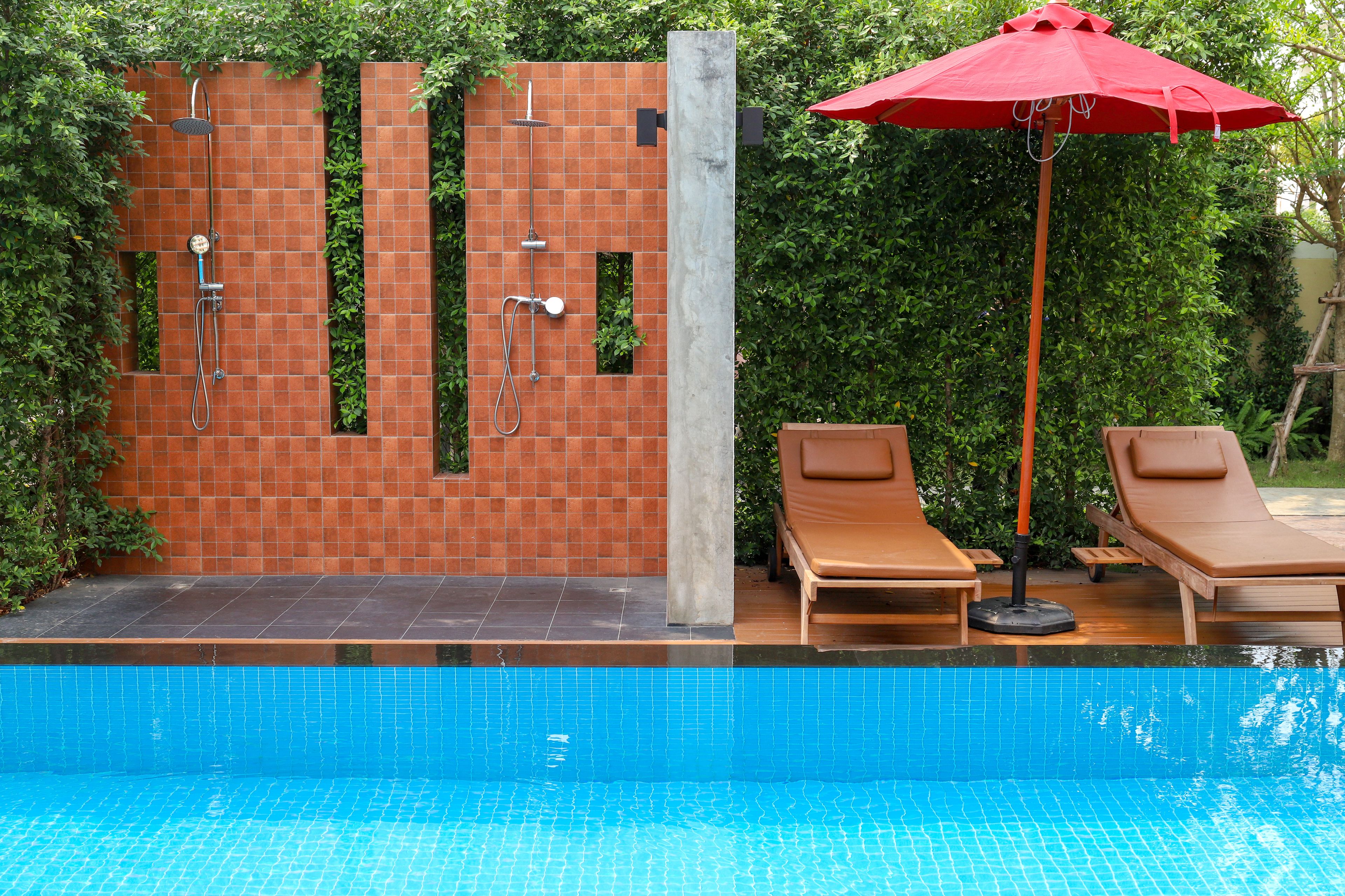 Una piscina con un muro de azulejos, una zona de ducha y dos sillas de descanso con una sombrilla roja.