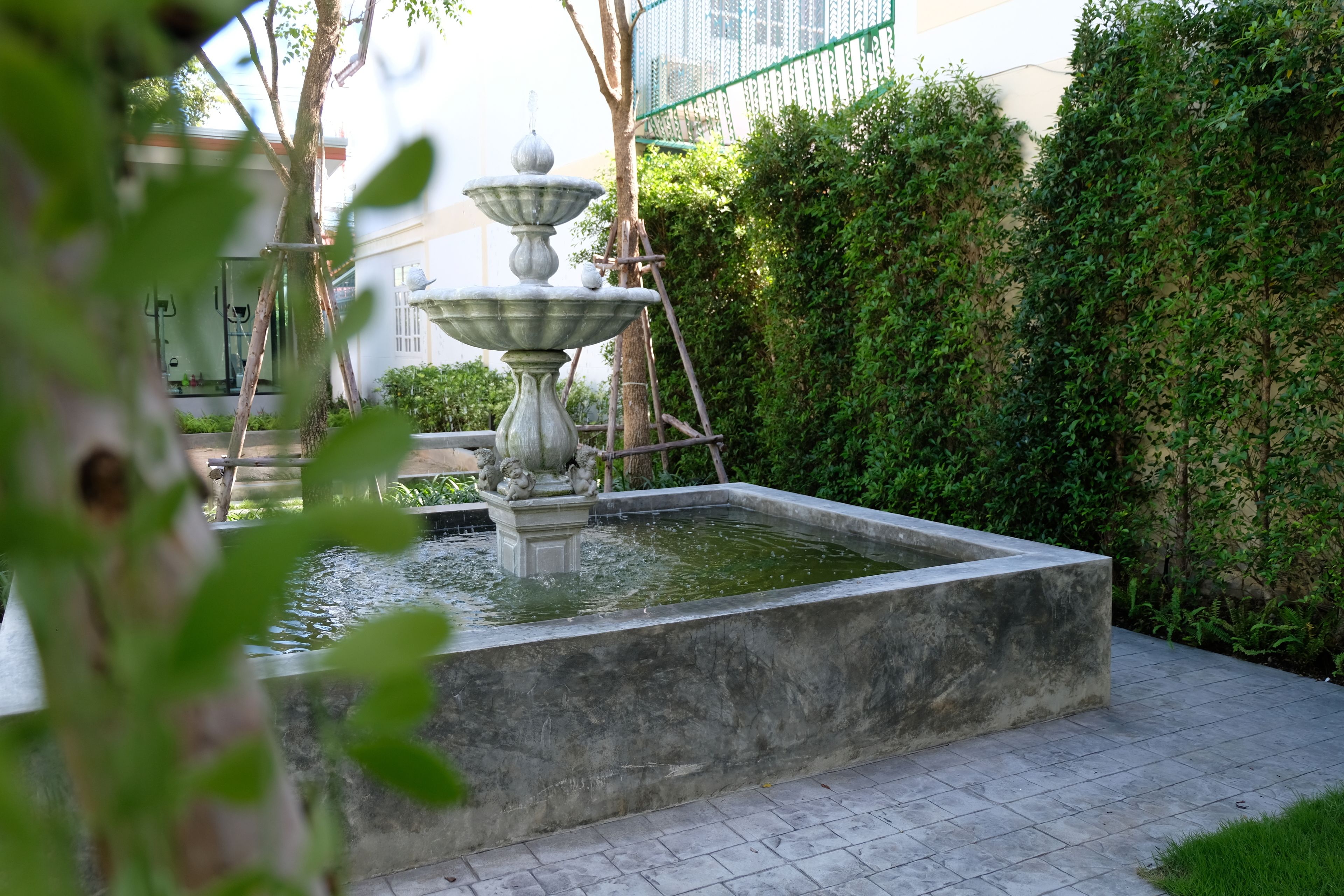 A fountain with three tiers and a rectangular basin, surrounded by a hedge and a building in the background.