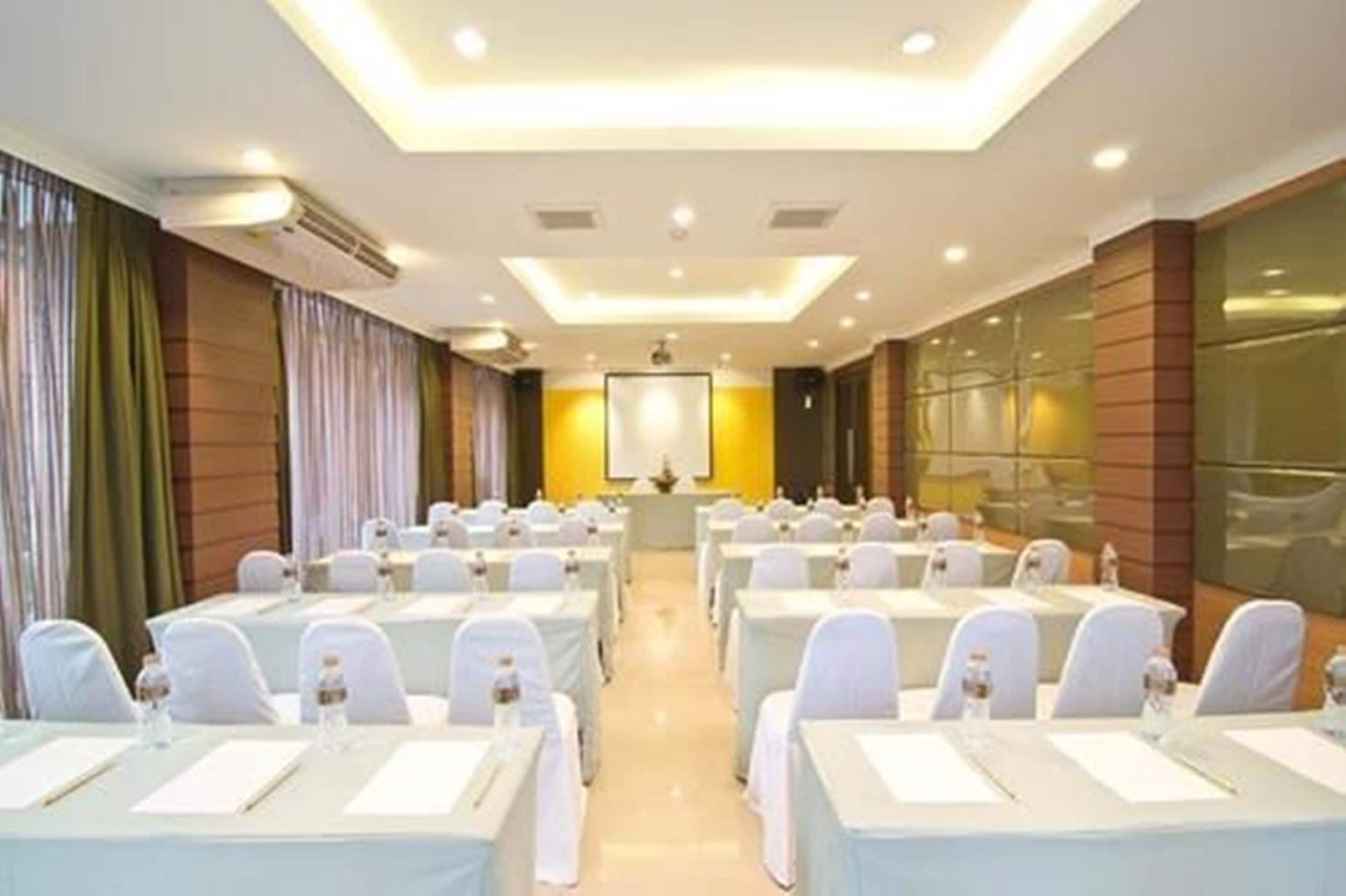 Une salle de conférence avec des rangées de tables, des chaises blanches et un écran de projection à l’avant.
