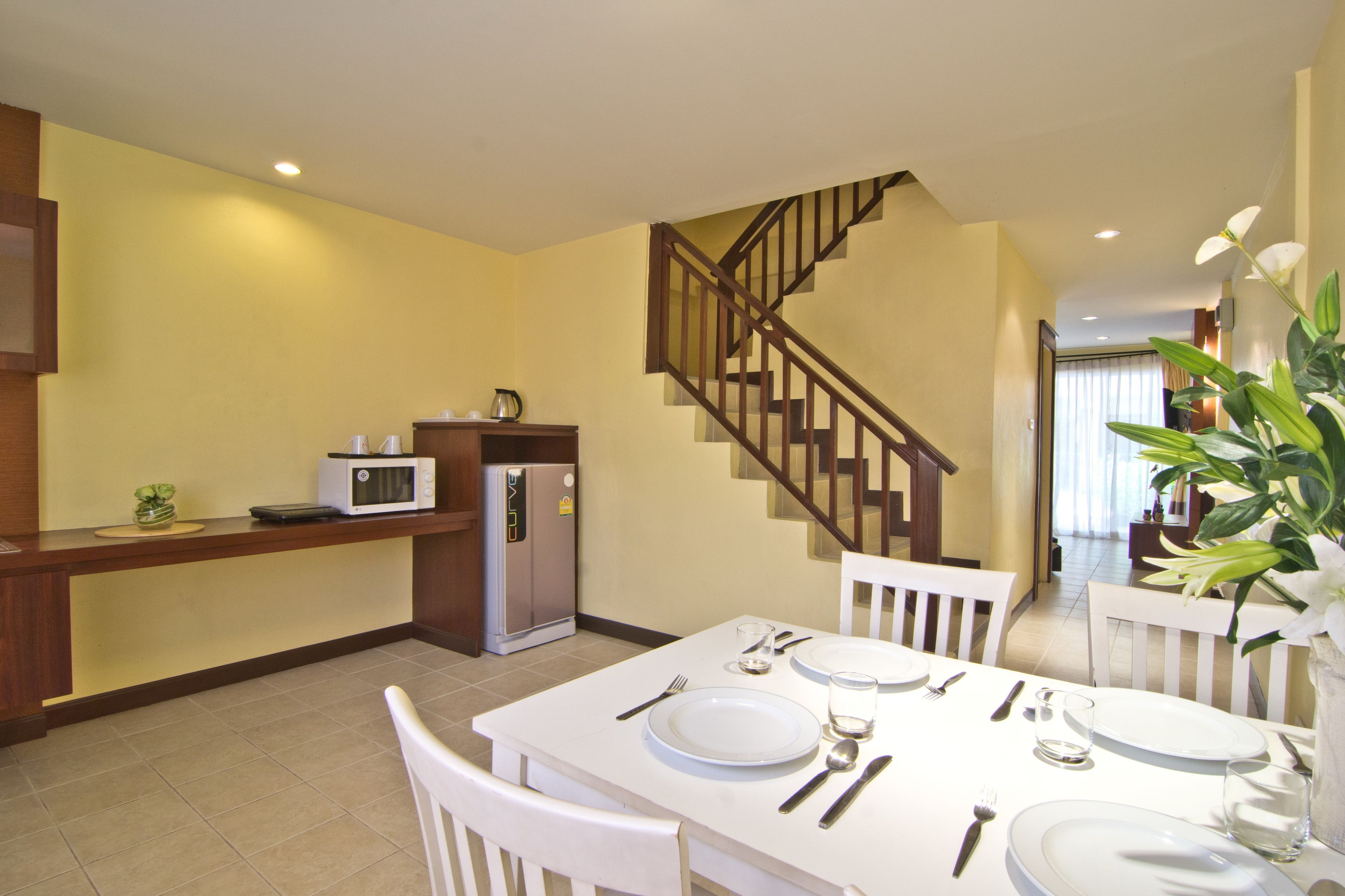 Un comedor con una mesa blanca dispuesta para cuatro, una cocina con microondas y refrigerador, y una escalera de madera.