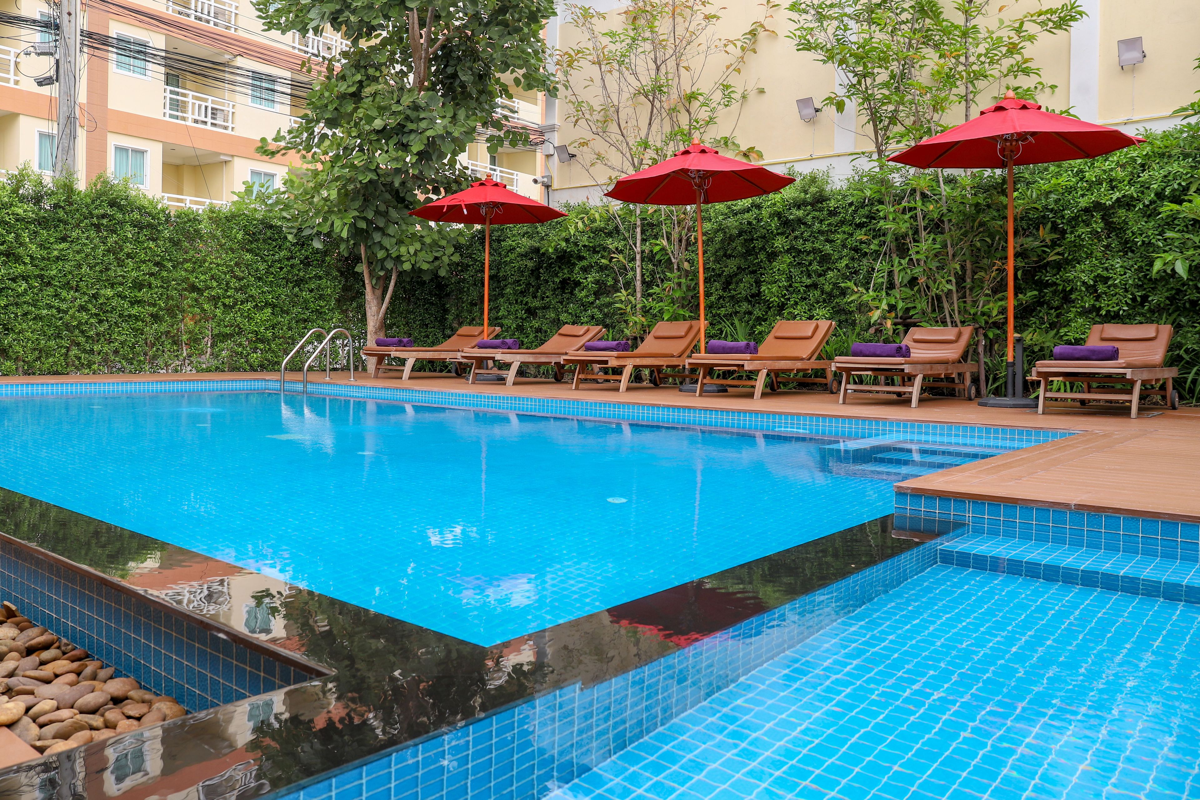 A swimming pool with lounge chairs and red umbrellas, surrounded by greenery and a building in the background.