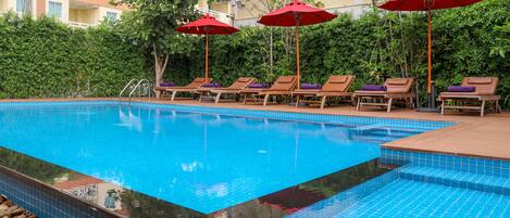 A swimming pool with lounge chairs and red umbrellas, surrounded by greenery and a building in the background.