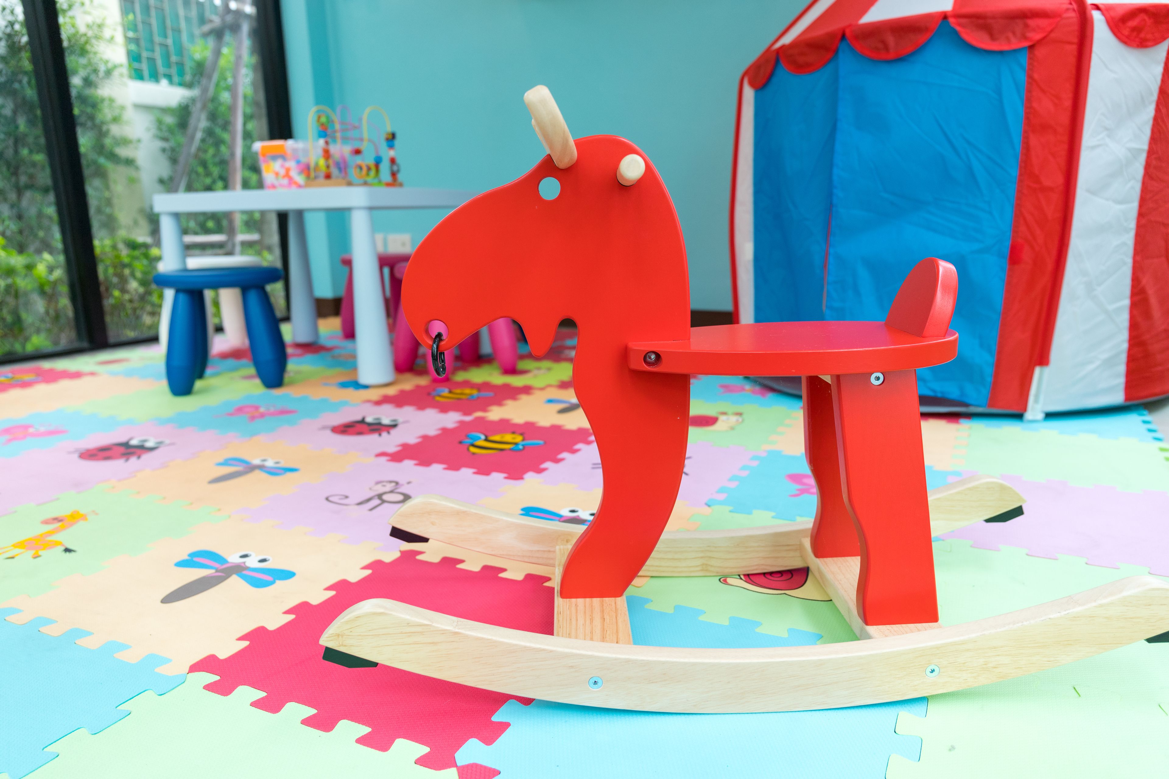 A rocking horse on a colorful play mat with a table and chairs in the background.