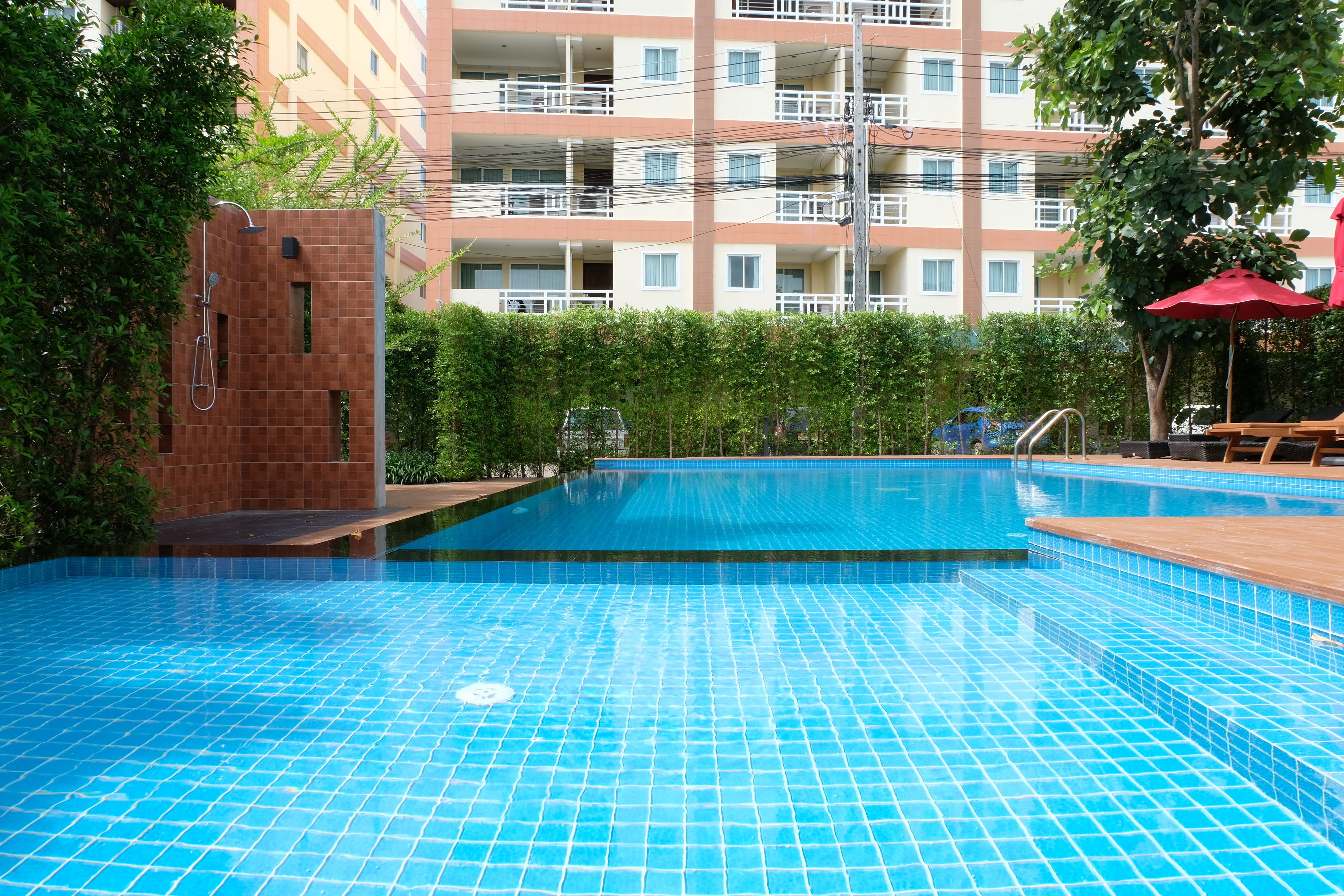A swimming pool with blue tiles, surrounded by greenery and a multi-story building in the background.