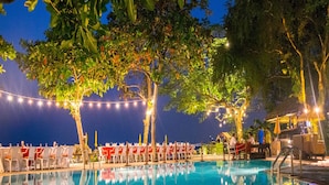 Un área de piscina nocturna con luces colgantes, sillones de descanso y una zona de bar.